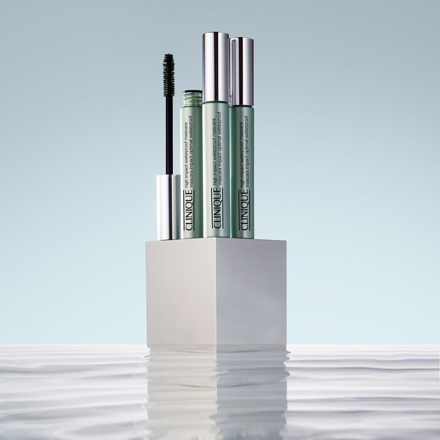 Trois tubes de mascara CLINIQUE High Impact Waterproof sur un piédestal blanc. Fond : eau avec reflets.