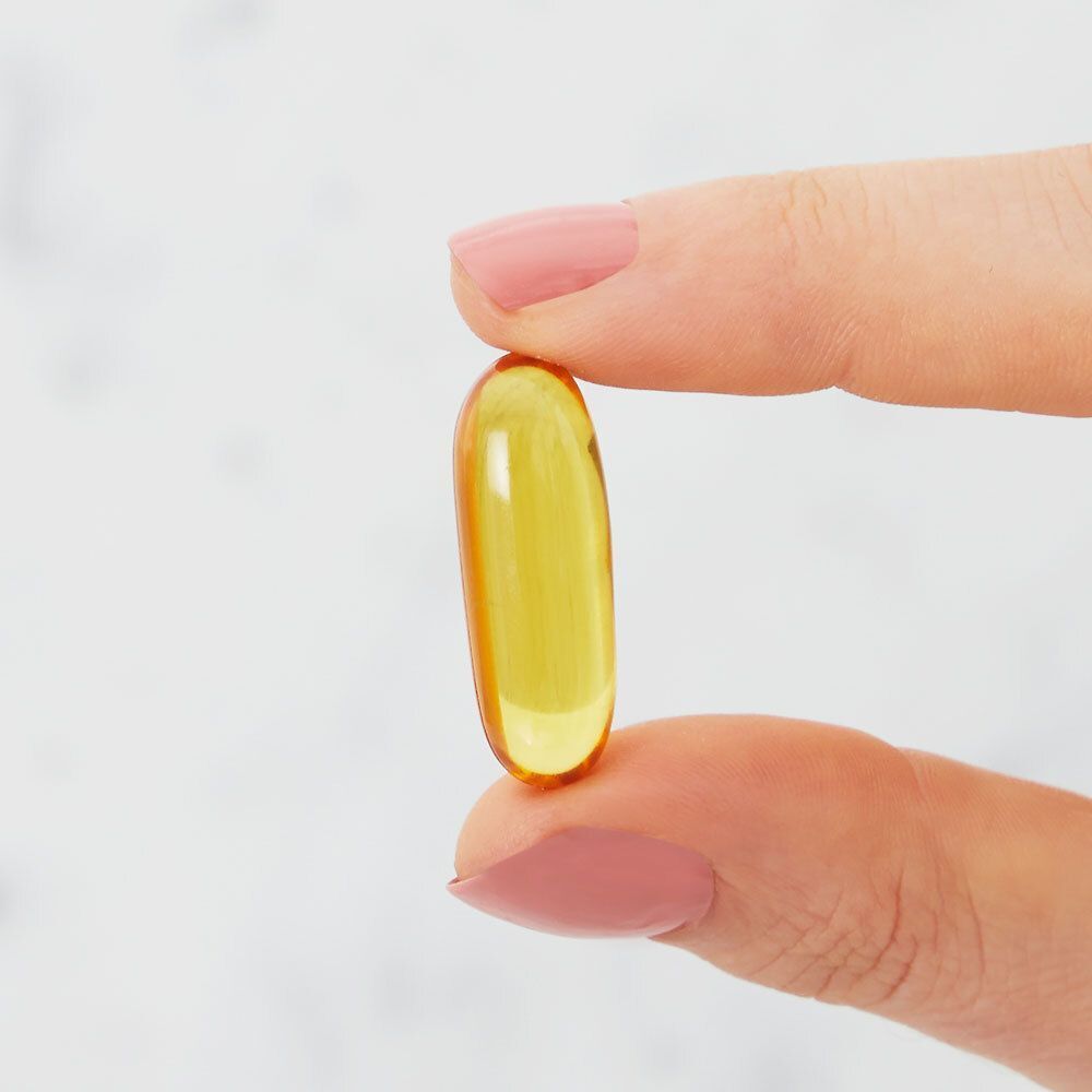 Une capsule ovale jaune tenue entre deux doigts. Peau claire et fond.