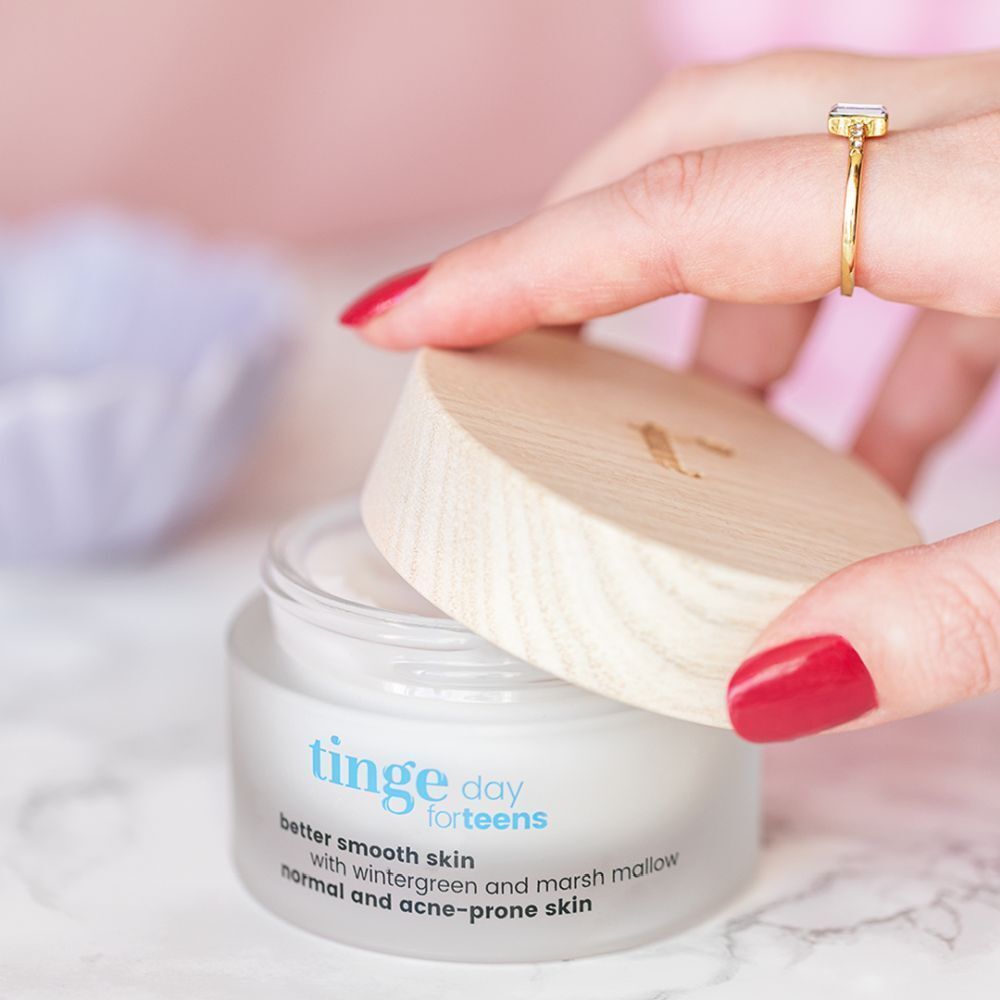 Un pot avec couvercle en bois clair, ouvert par une main. Inscription: Tinge day for teens, better smooth skin, avec gaulthérie et guimauve, peaux normales et acnéiques.
