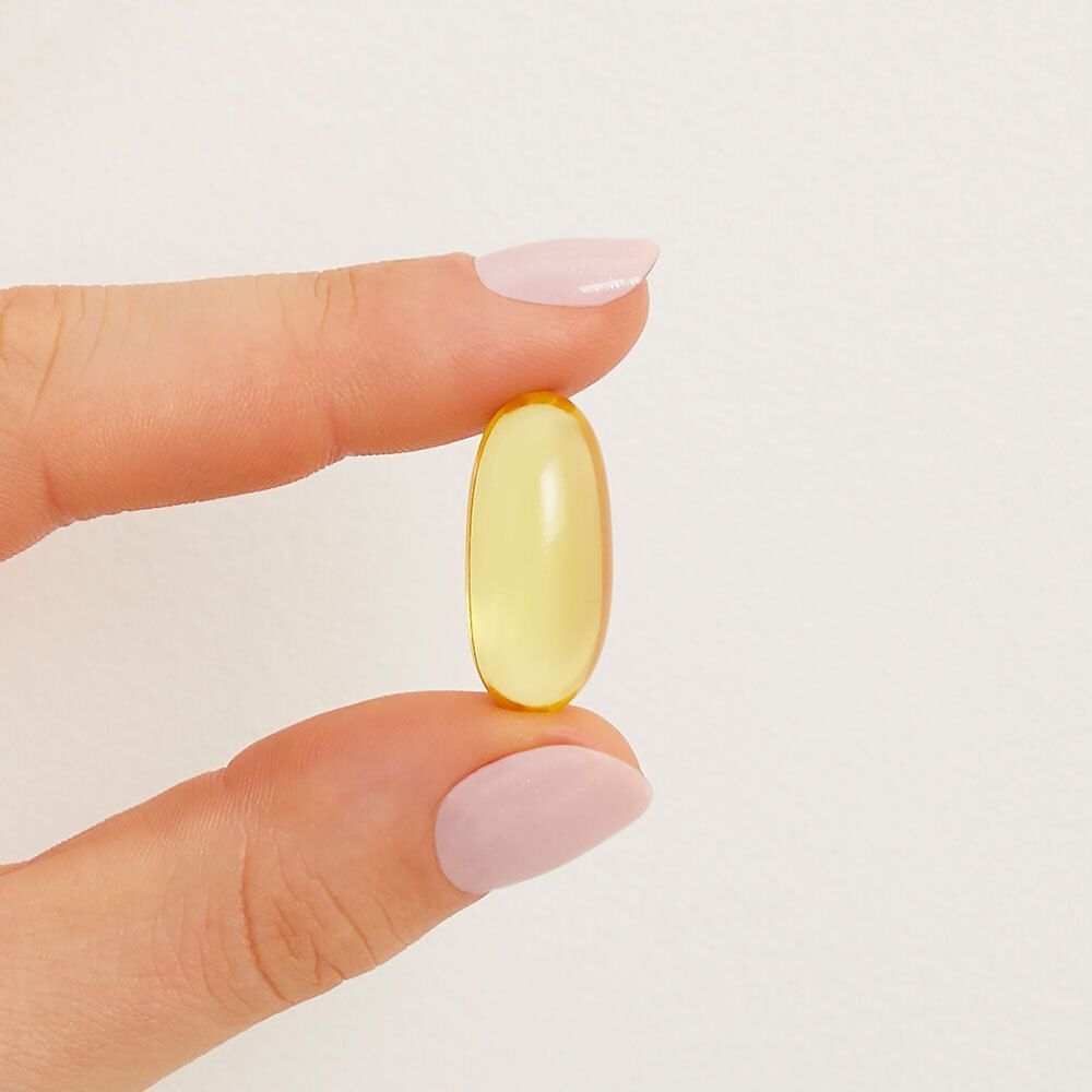 Une main tient une gélule ovale jaune entre le pouce et l'index sur fond blanc.