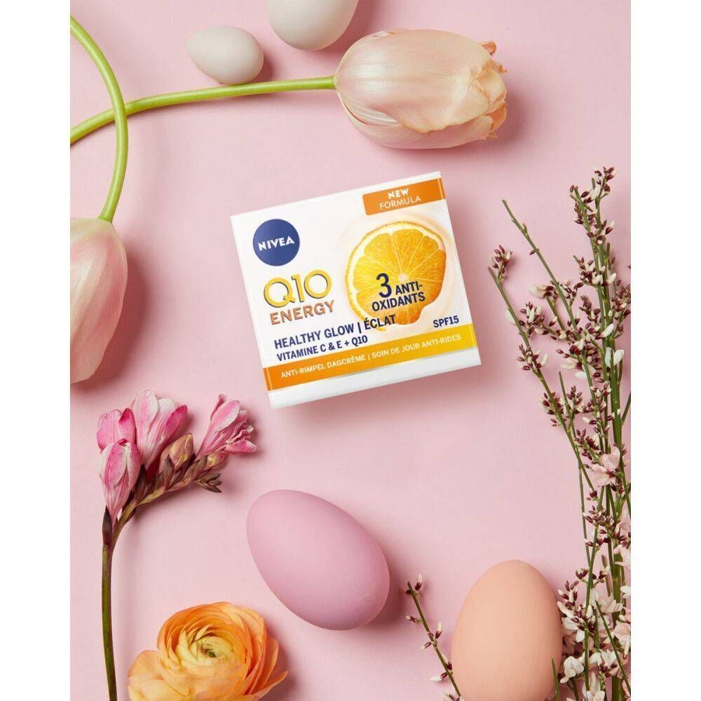 Emballage blanc avec logo NIVEA et inscription jaune Q10 ENERGY, entouré de fleurs et d'œufs sur fond rose. Texte Healthy Glow, SPF15.