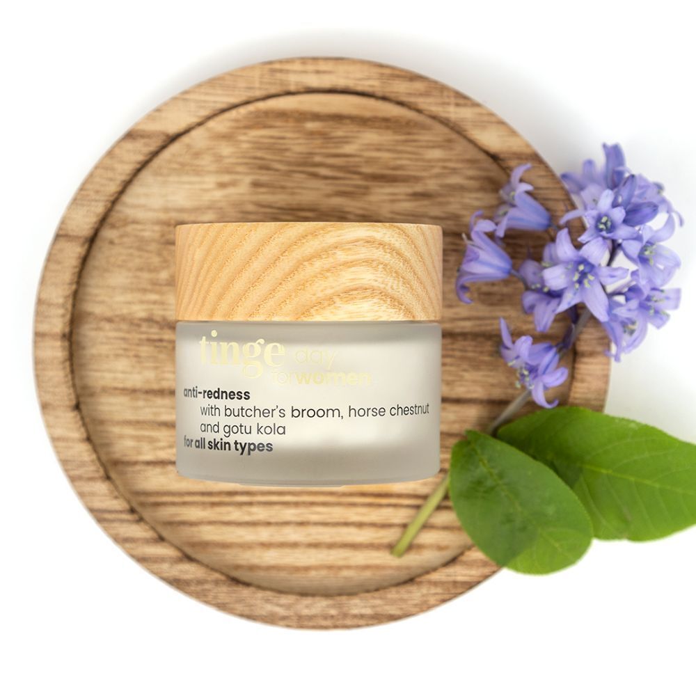 Pot de crème anti-rougeurs avec couvercle en bois, sur dessous de plat en bois. Fleurs bleues et feuilles à côté. Texte: anti-redness, for all skin types.