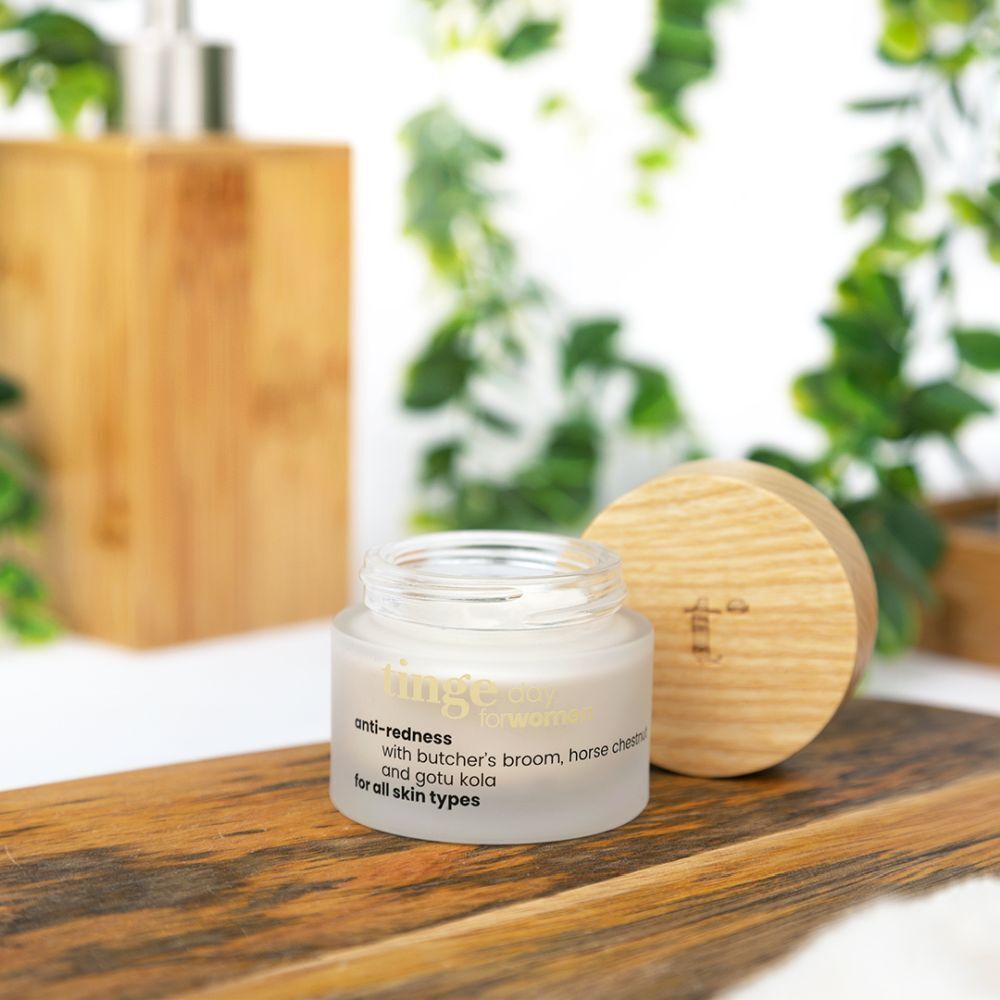 Pot de crème anti-rougeurs avec couvercle en bois, sur planche en bois. Distributeur de savon en bois et plantes vertes à côté. Texte: anti-redness, for all skin types.