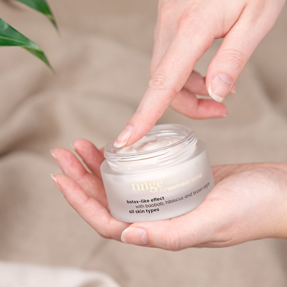 Mains tenant un pot de Crème de Jour Intense. Verre blanc, couvercle en bois. Inscription : Tinge, botox-like effect, pour tous types de peau.
