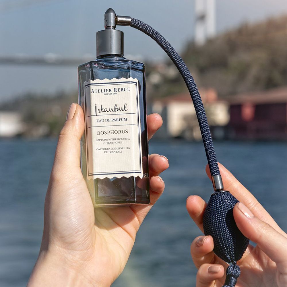 Mains tenant un flacon bleu foncé avec vaporisateur. Inscription: Istanbul Bosphorus Eau De Parfum. Fond: eau et pont.