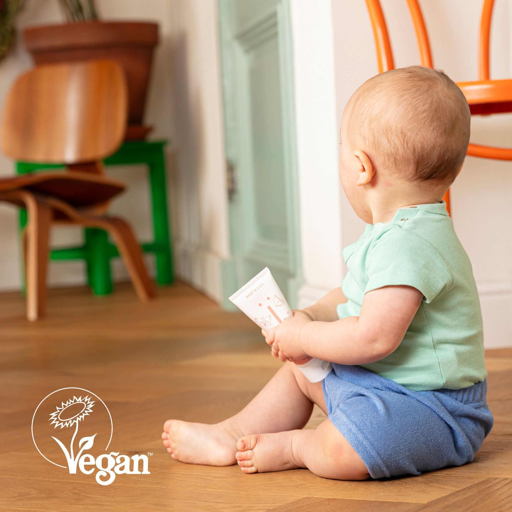 Bébé assis par terre tenant un tube de crème au zinc NAÏF, sceau Vegan.