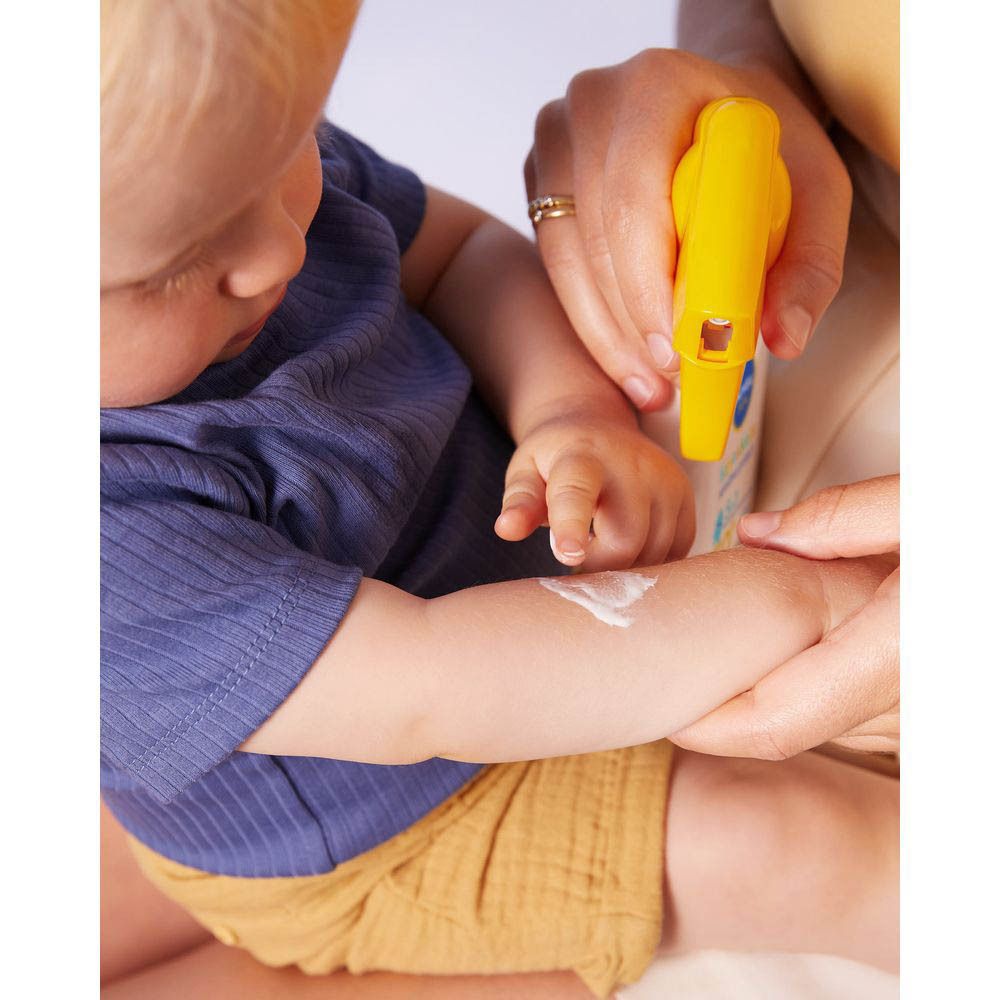 Bébé, bras vaporisé de crème solaire. Pantalon jaune, vêtements bleus.