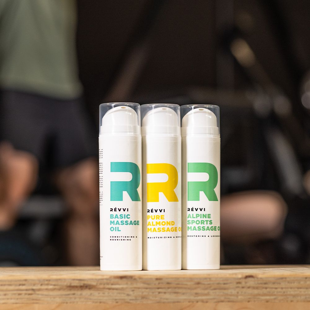 Trois flacons blancs avec logos colorés. Inscription : RÉVVI, BASIC MASSAGE OIL, PURE ALMOND MASSAGE, ALPINE SPORTS MASSAGE OIL.