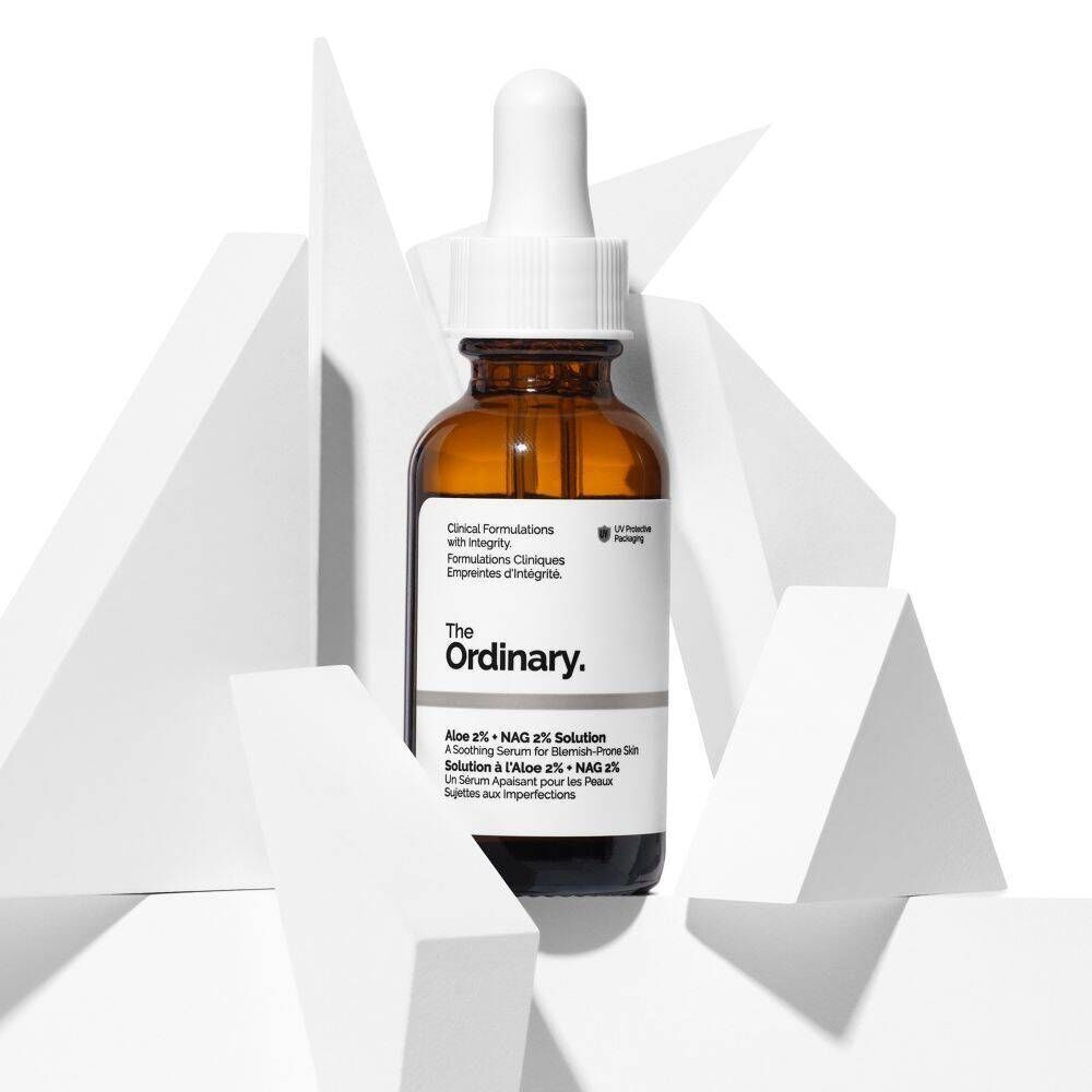 Un flacon de 'The Ordinary Aloe 2% + NAG 2% Solution' avec pipette, entouré de formes géométriques blanches.