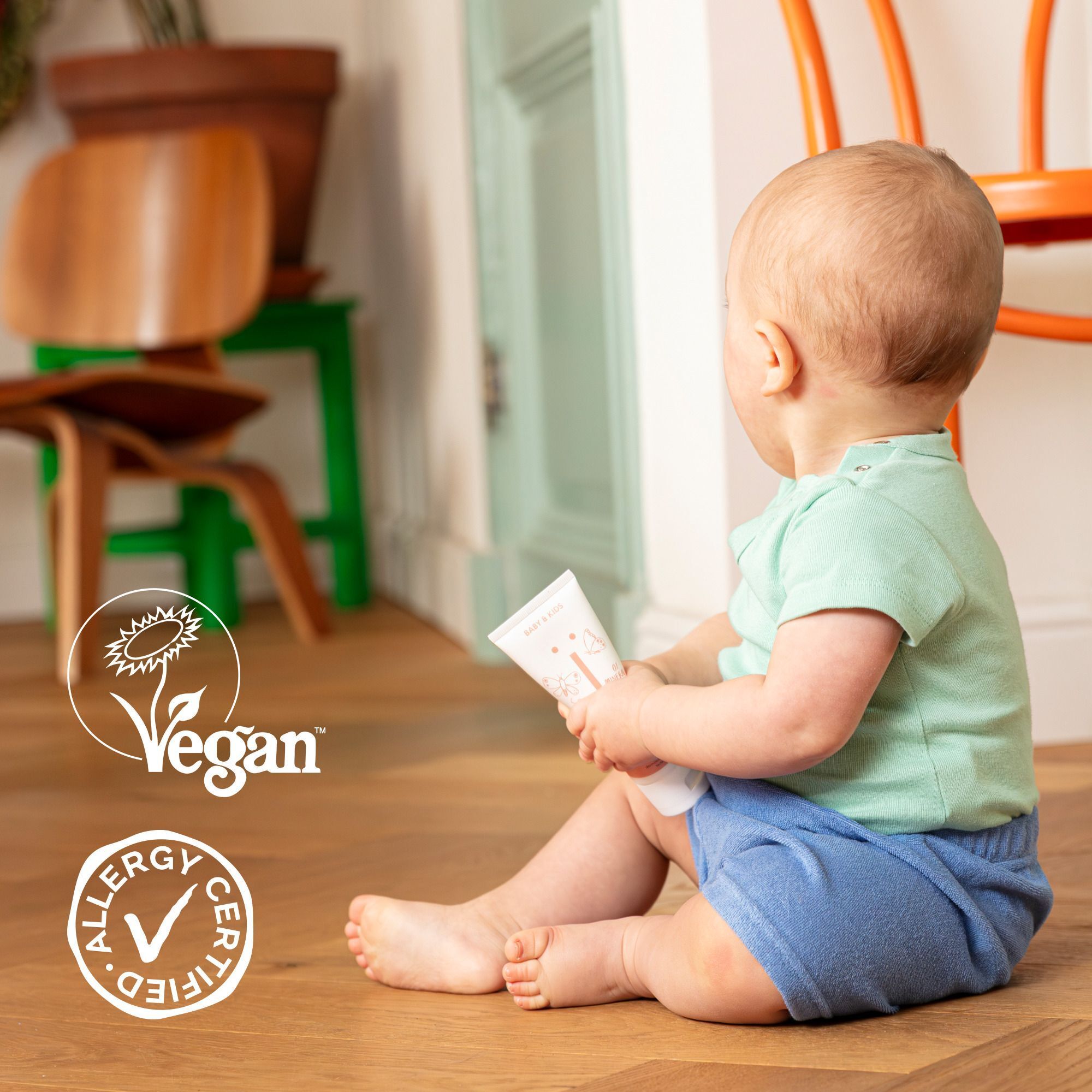 Un bébé assis par terre tenant un flacon de Naïf Baby Powder. Logos Vegan et Allergy Certified.