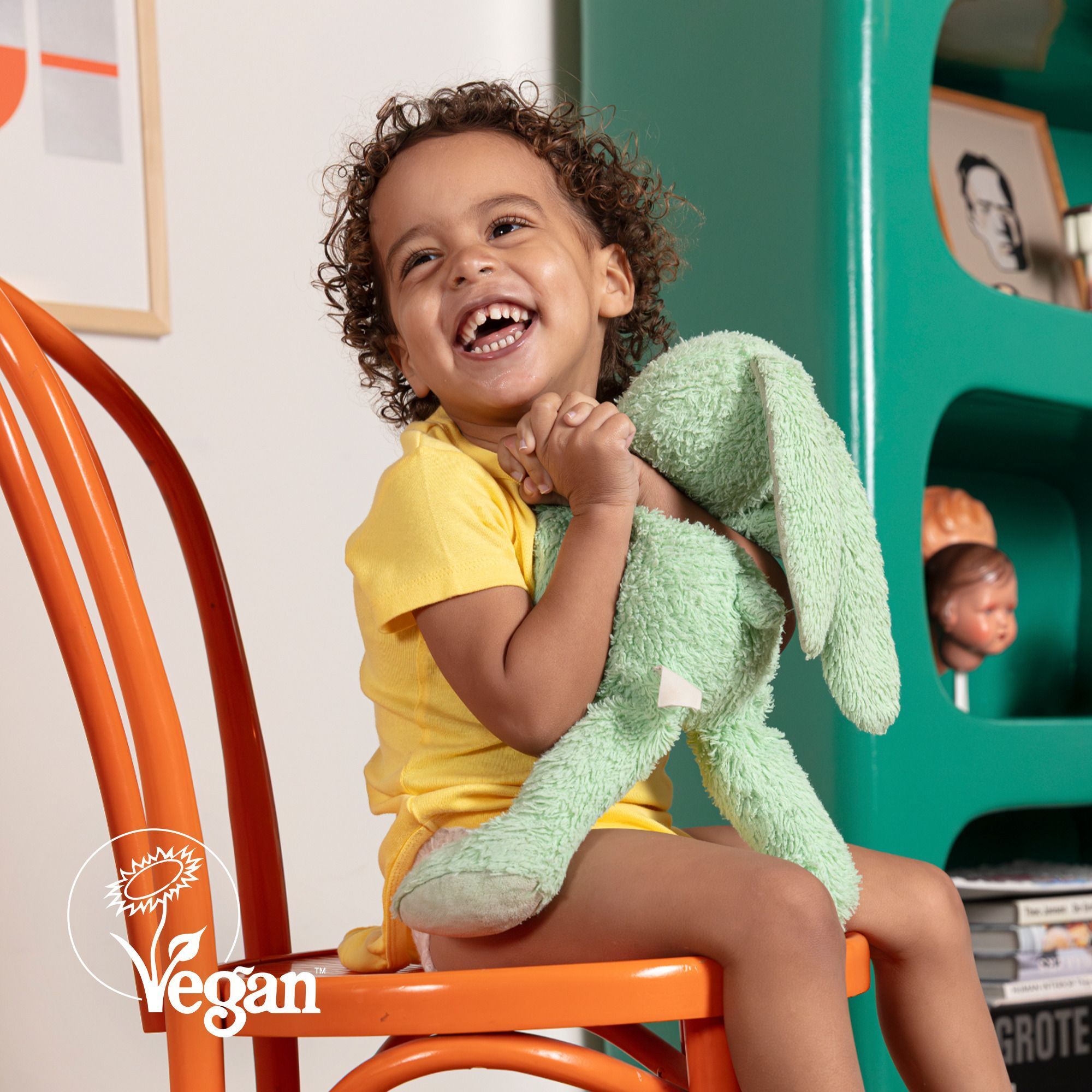 Un bébé souriant avec un lapin en peluche sur une chaise, avec un sceau Vegan.