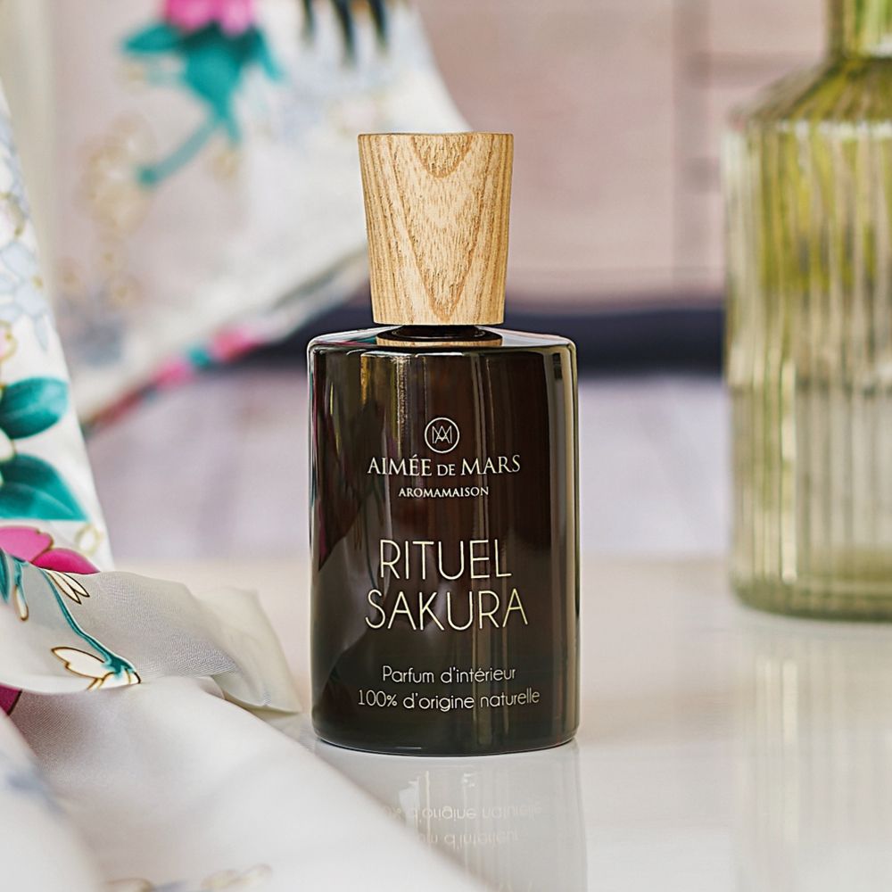 Flacon en verre brun avec bouchon en bois. Inscription: RITUEL SAKURA, Parfum d'intérieur, 100% d'origine naturelle. Sur une table.