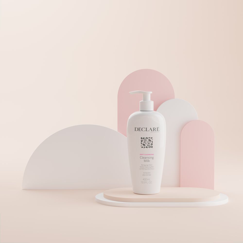 Flacon blanc DECLARÉ Cleansing Milk sur socle. Formes de fond roses et blanches.