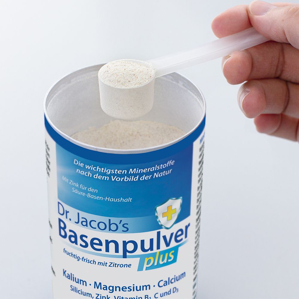Main tenant une cuillère doseuse de poudre au-dessus d'une boîte de Dr. Jacob's Basenpulver plus. Texte : Basenpulver, Formule alcalinisante plus.