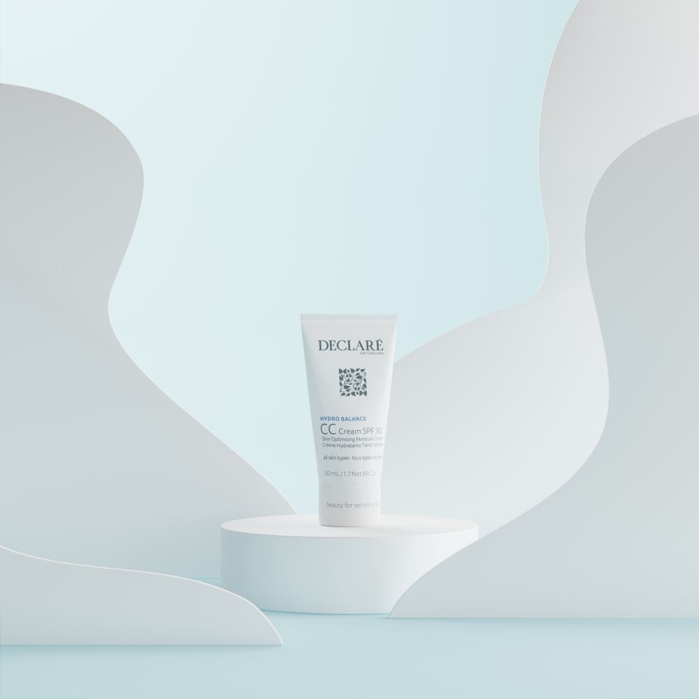 Tube blanc DECLARÉ CC Cream SPF 30 sur un piédestal. Fond avec formes blanches ondulées.