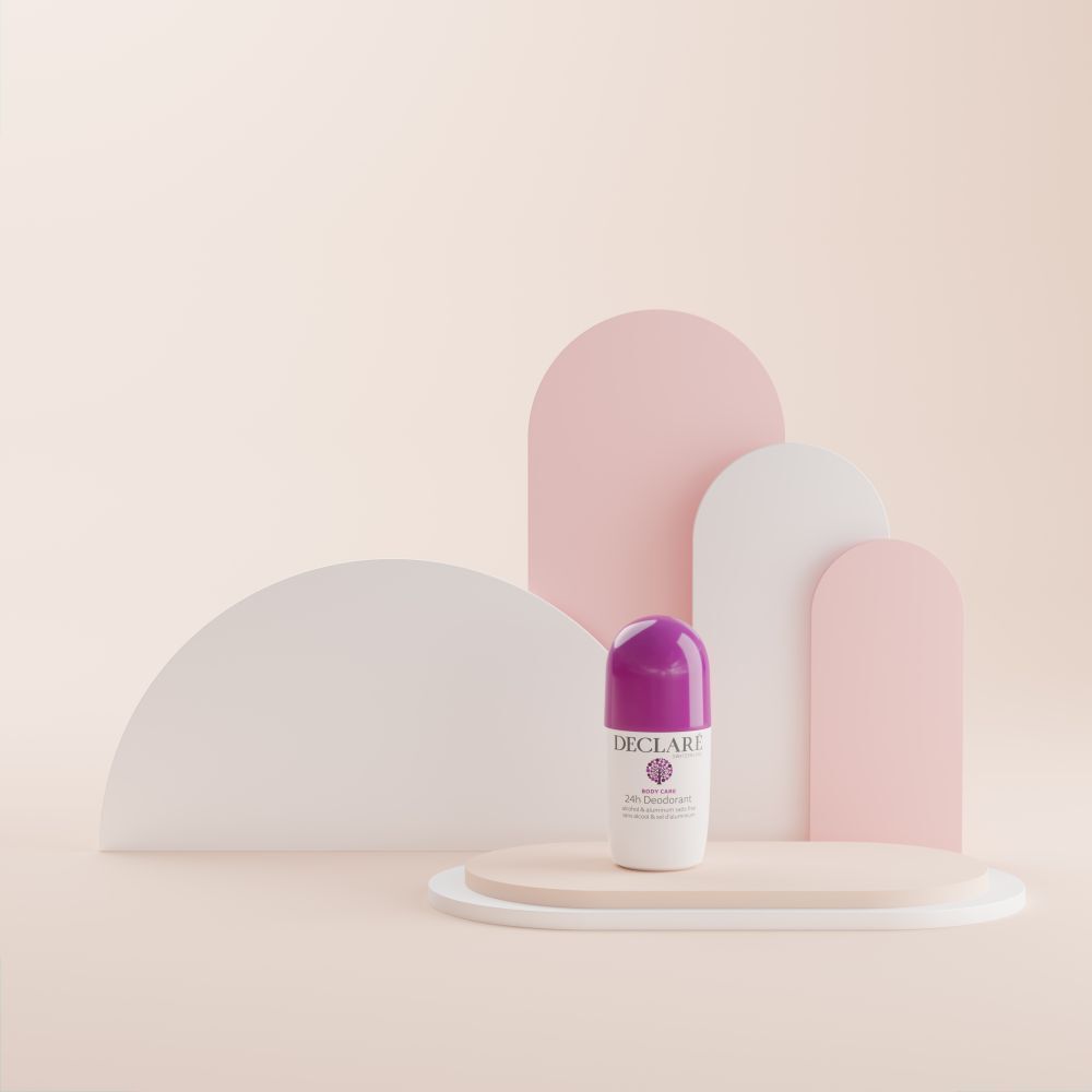 Déodorant roll-on sur socle. Boîtier blanc, bouchon violet. DECLARÉ, 24h Deodorant. Fond: formes roses et blanches incurvées.