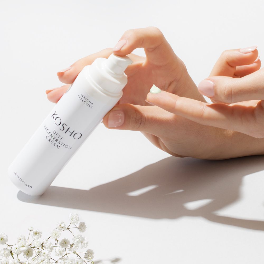 Main tenant un flacon blanc de KOSHO Deep Regeneration Cream. Crème appliquée sur les doigts. Fleurs blanches.