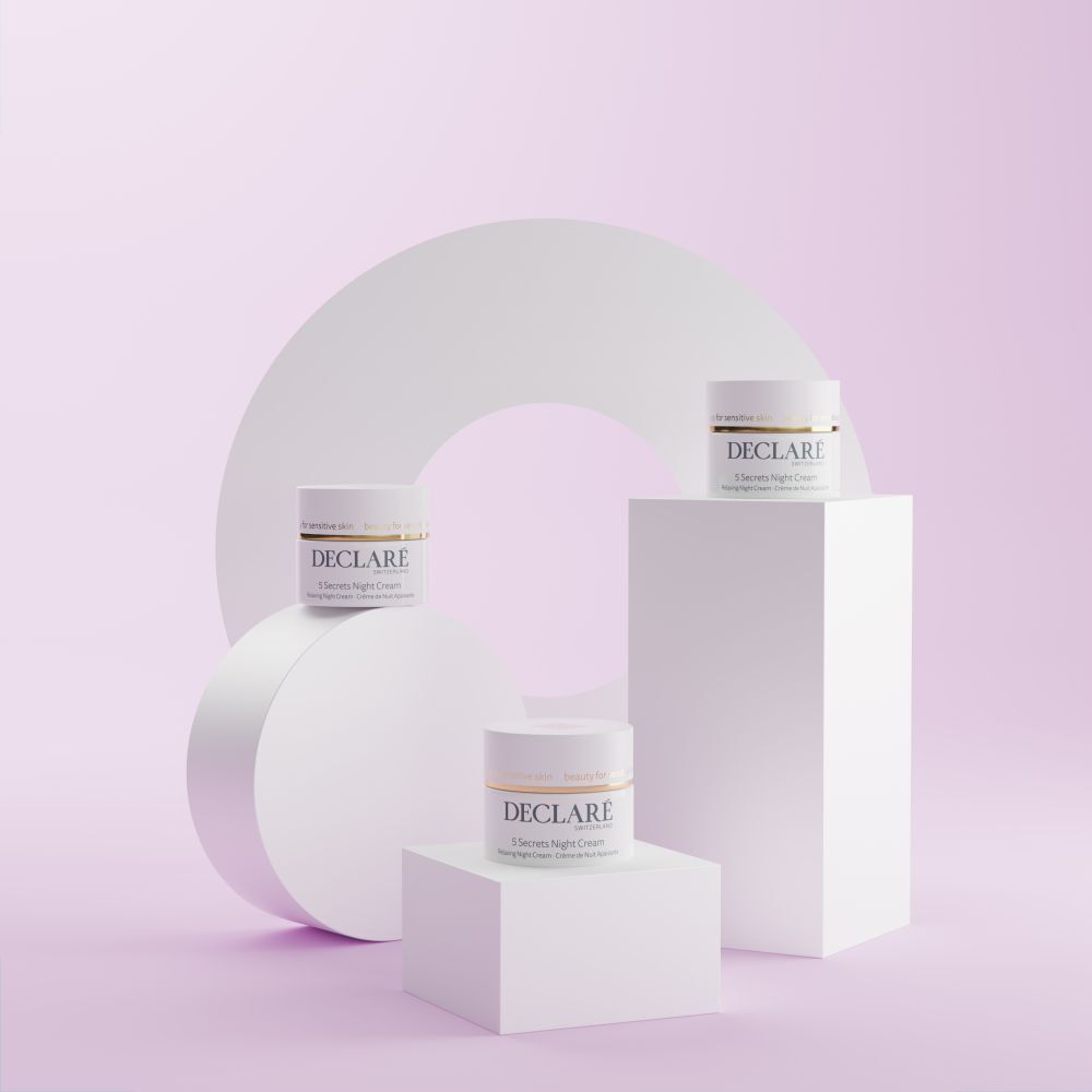 Trois pots blancs DECLARÉ 5 Secrets Night Cream sur socles. Fond: formes géométriques.