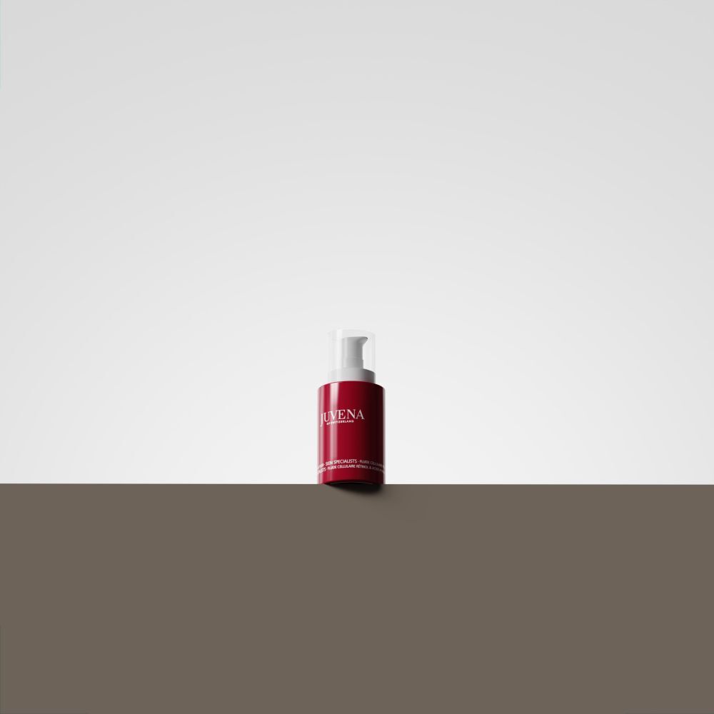 Flacon rouge avec bouchon blanc. Inscription: JUVENA. Posé sur une surface marron devant un fond blanc.