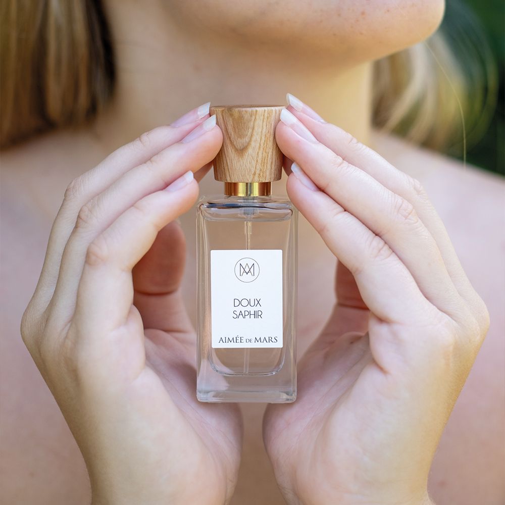 Mains tenant un flacon de parfum avec bouchon en bois. Étiquette: "Doux Saphir" et "Aimée de Mars".