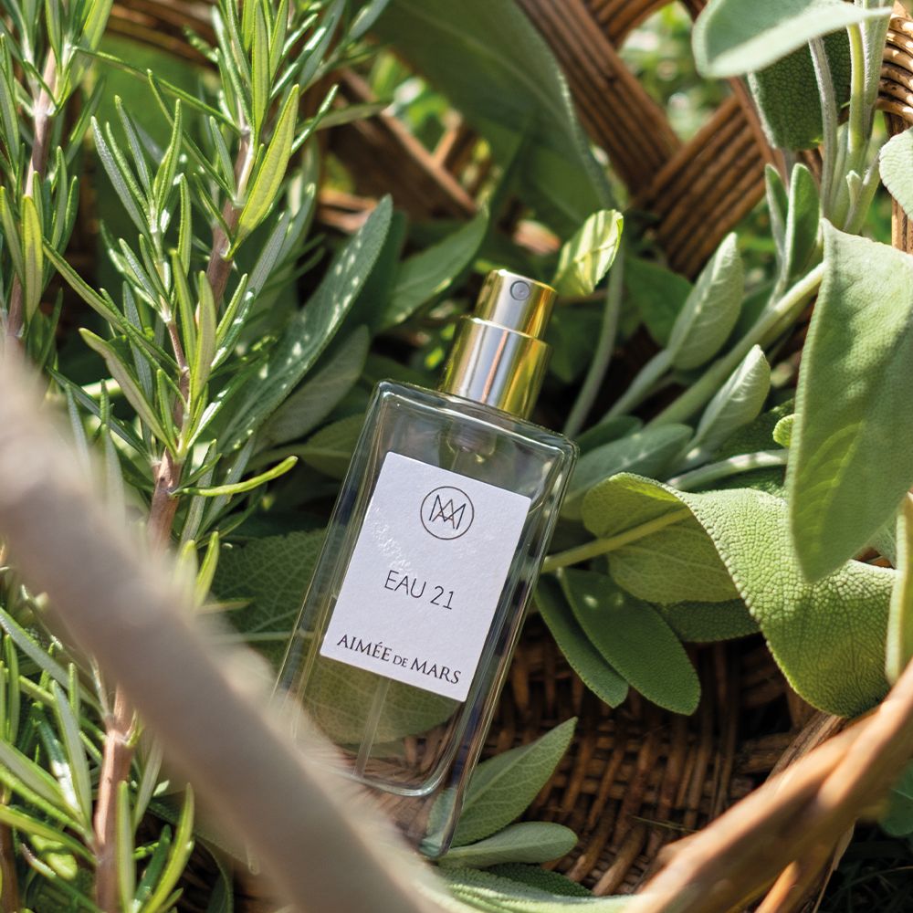 Flacon de parfum entouré d'herbes. Étiquette avec logo, "EAU 21" et "Aimée de Mars".