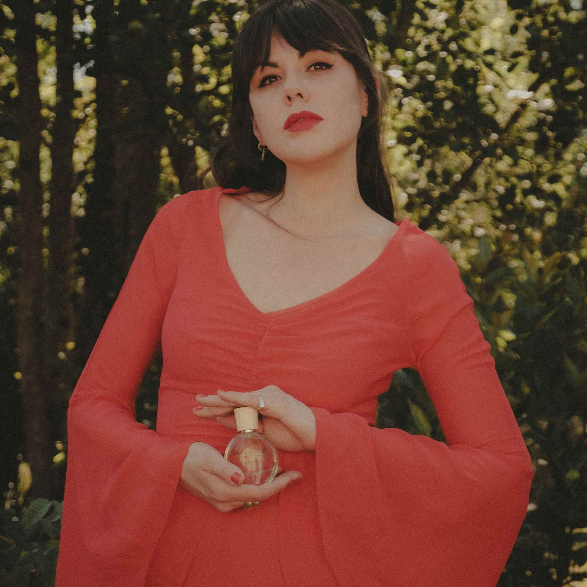 Femme tenant un flacon. Elle porte une robe rouge. Le flacon est visible.