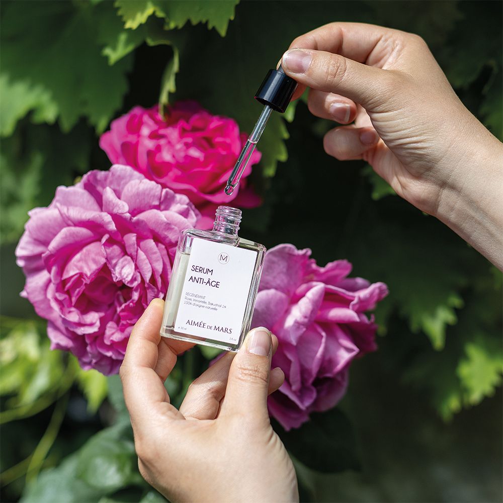 Mains tenant flacon et pipette au-dessus de roses. Flacon "SERUM ANTI-ÂGE" et "AIMÉE DE MARS".