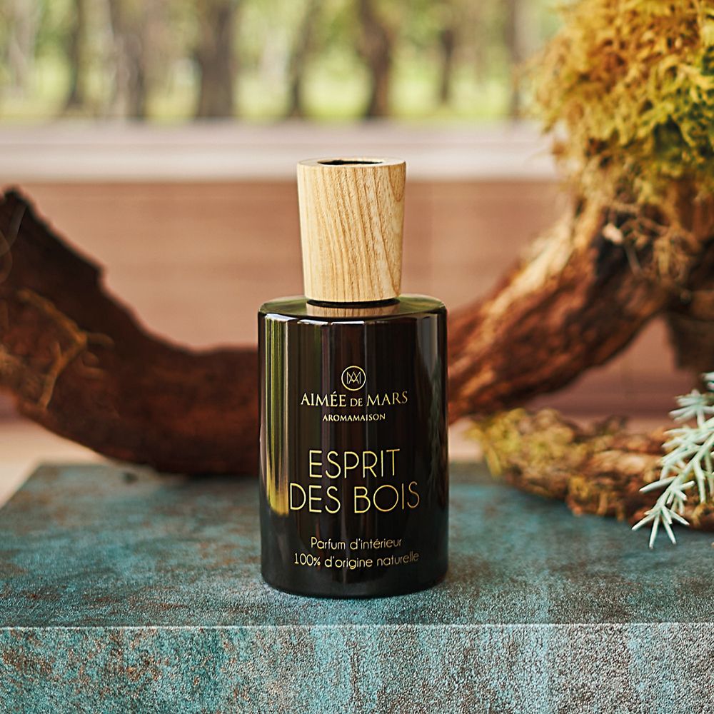 Flacon en verre brun avec bouchon en bois, devant une branche et des plantes. Inscription: Esprit des Bois, Parfum d'intérieur.