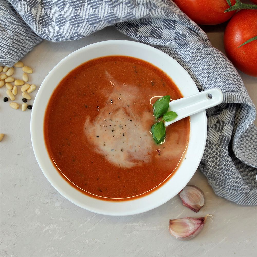 Soupe de tomates dans un bol blanc avec une cuillère. Garnie de basilic. Sur fond gris avec tomates, ail et pignons de pin.