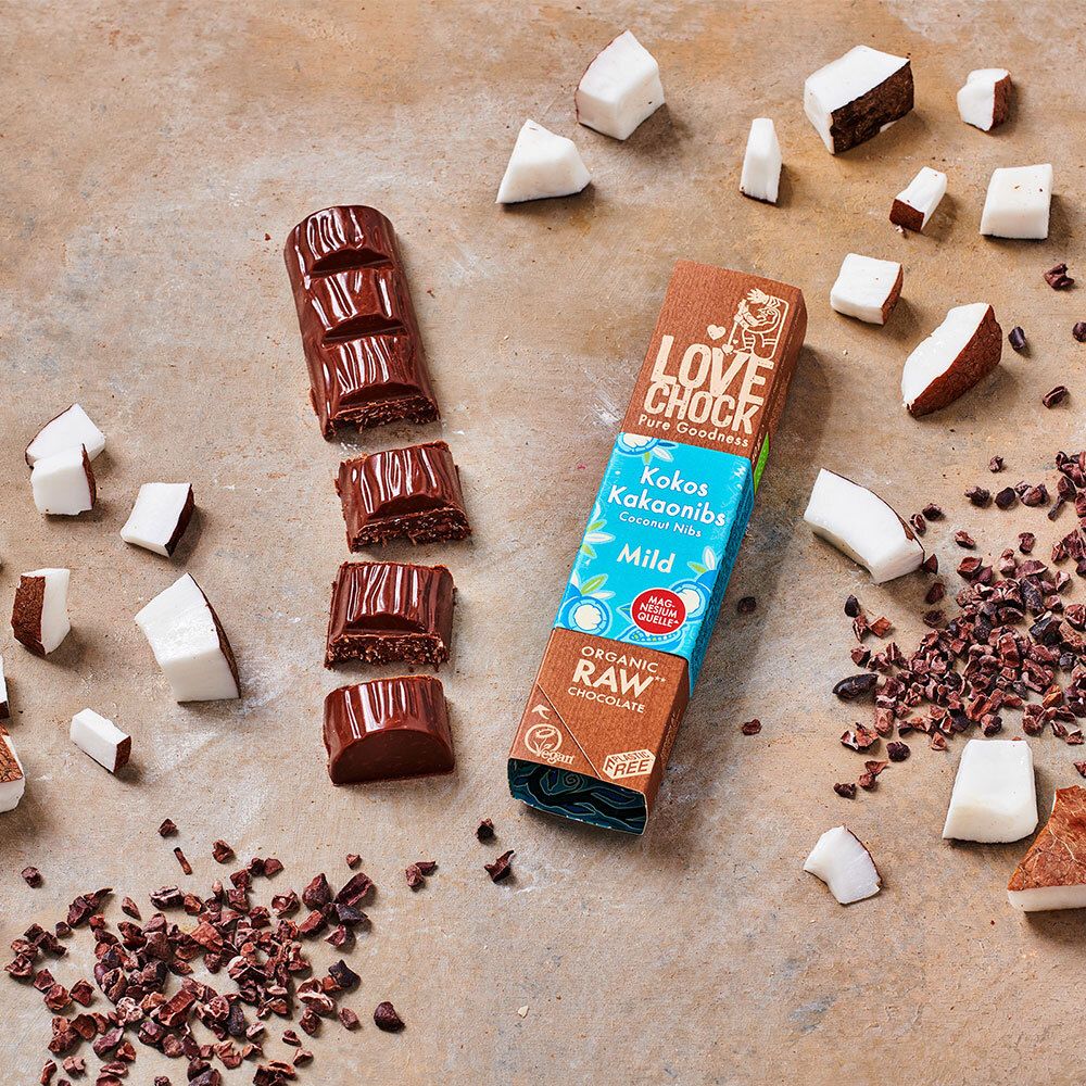 Barre LOVECHOCK Kokos Kakaonibs Mild, partiellement cassée, entourée de morceaux de noix de coco et de nibs de cacao.