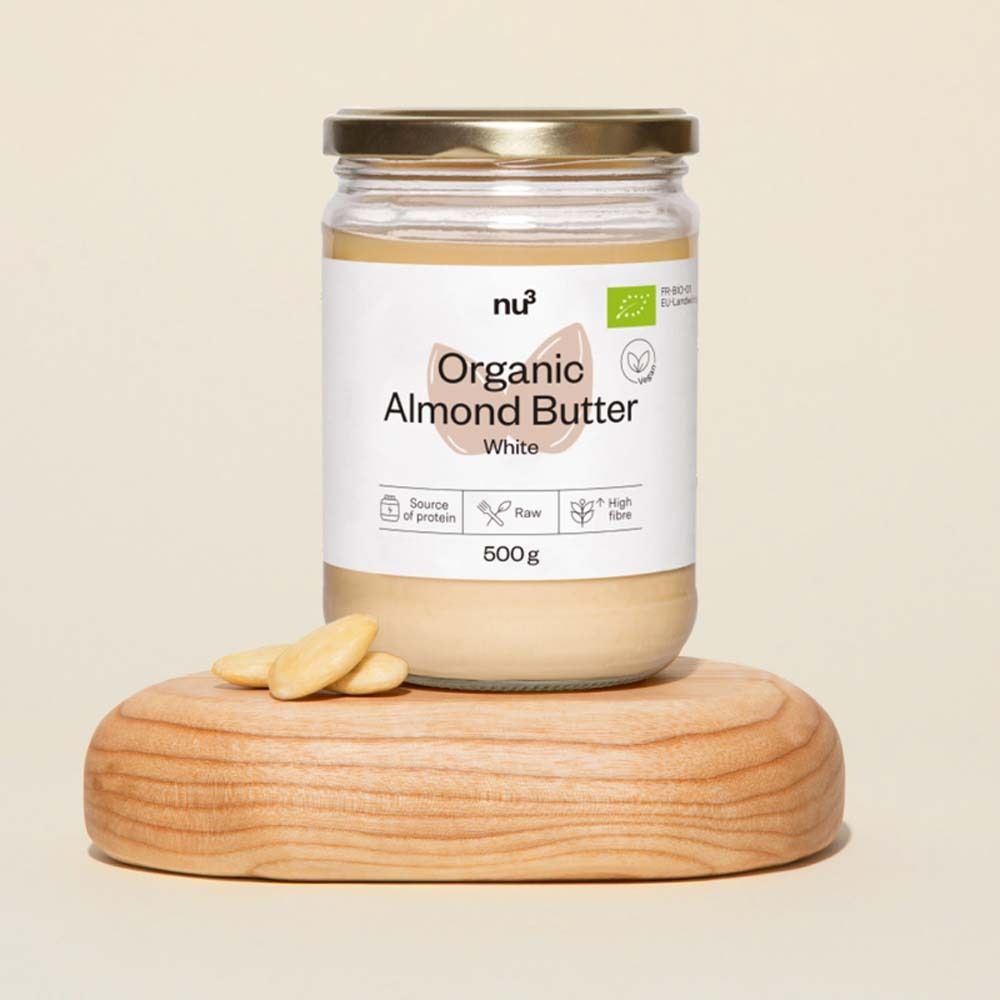 Pot de beurre d'amandes blanches nu3 Bio, avec couvercle doré et étiquette blanche. Inscription : Organic Almond Butter White, 500g. Éclats d'amandes sur planche.
