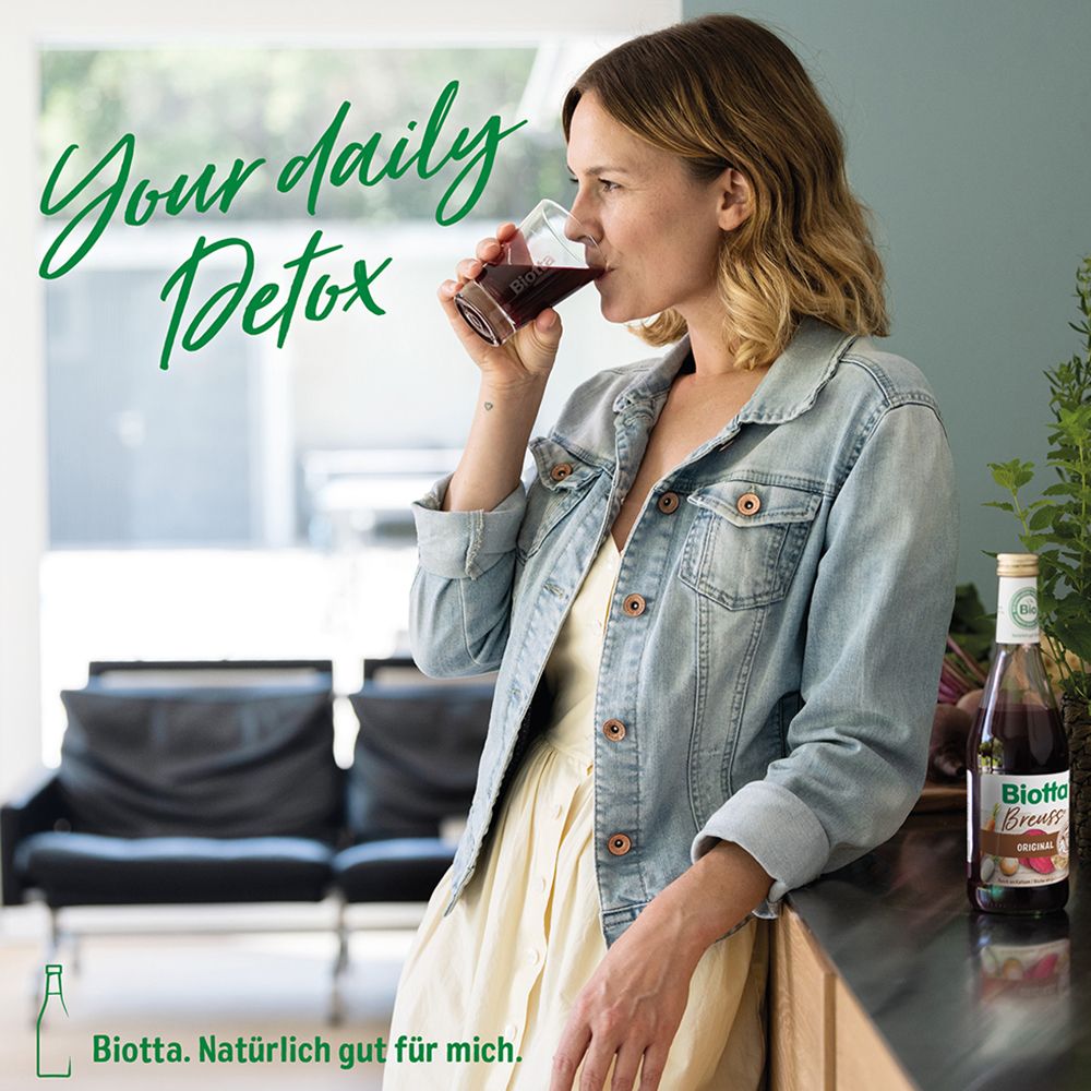 Femme buvant du jus dans un verre. Bouteille Biotta Breuss Original en arrière-plan. Texte : Your daily Detox.
