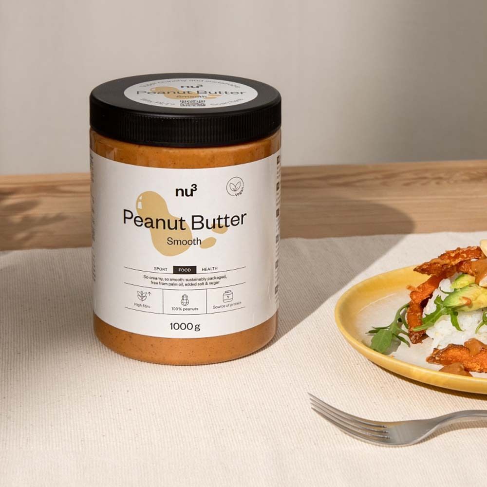Un pot de beurre de cacahuète nu3 smooth. Sur une table, à côté d'une assiette de nourriture. Couvercle noir, étiquette blanche.