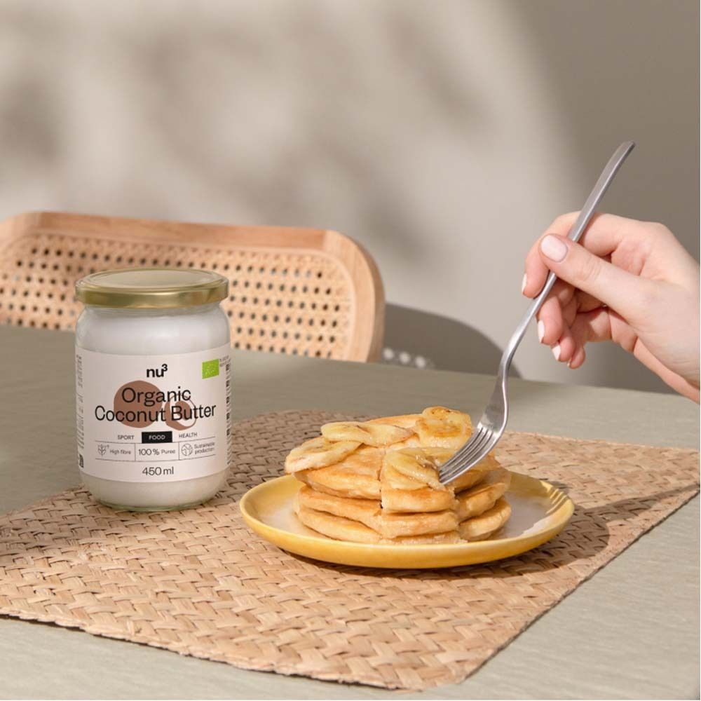 Pot de beurre de coco bio à côté de crêpes sur une assiette jaune. Personne tient une fourchette. Inscription : Organic Coconut Butter, 450ml.
