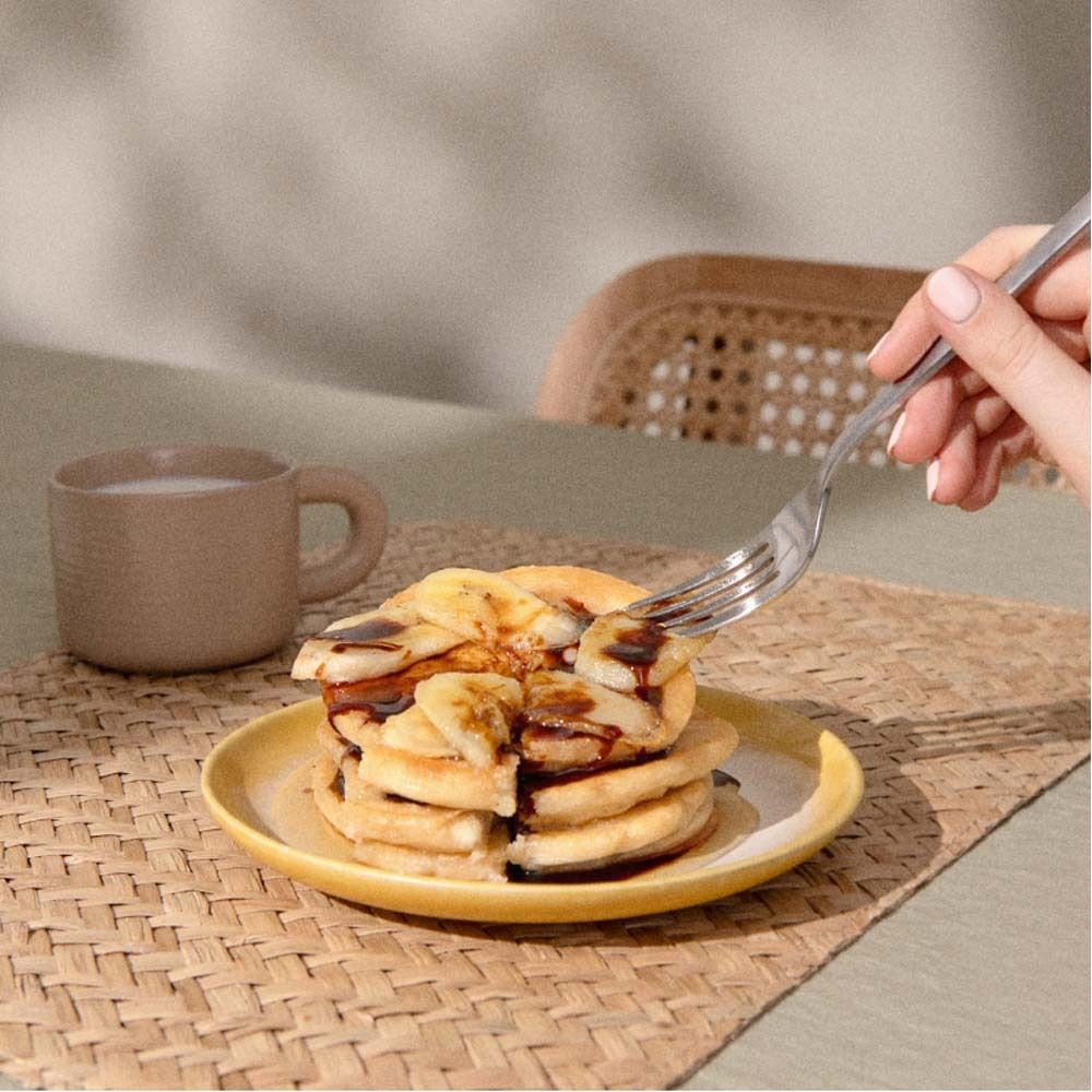 Crêpes avec tranches de banane sur une assiette. Une main tient une fourchette. Une tasse en arrière-plan.