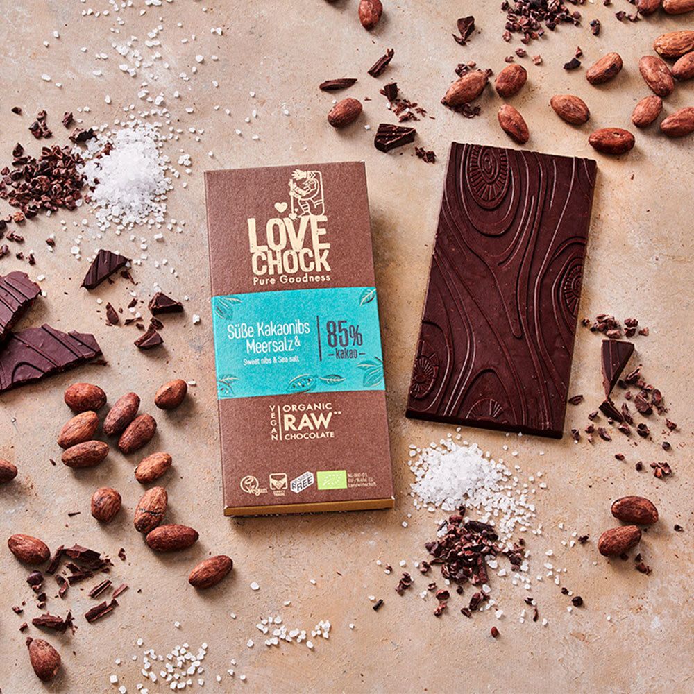 Tablette de chocolat LOVECHOCK à côté d'une tablette cassée. Fèves de cacao et sel marin dispersés. Inscription : Süße Kakaonibs & Meersalz, 85% Kakao.