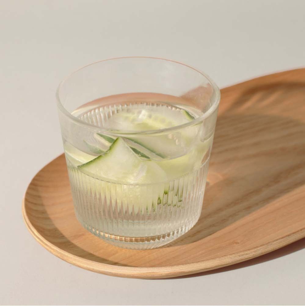 Verre avec boisson et tranches de concombre, sur plateau en bois. Fond : clair.