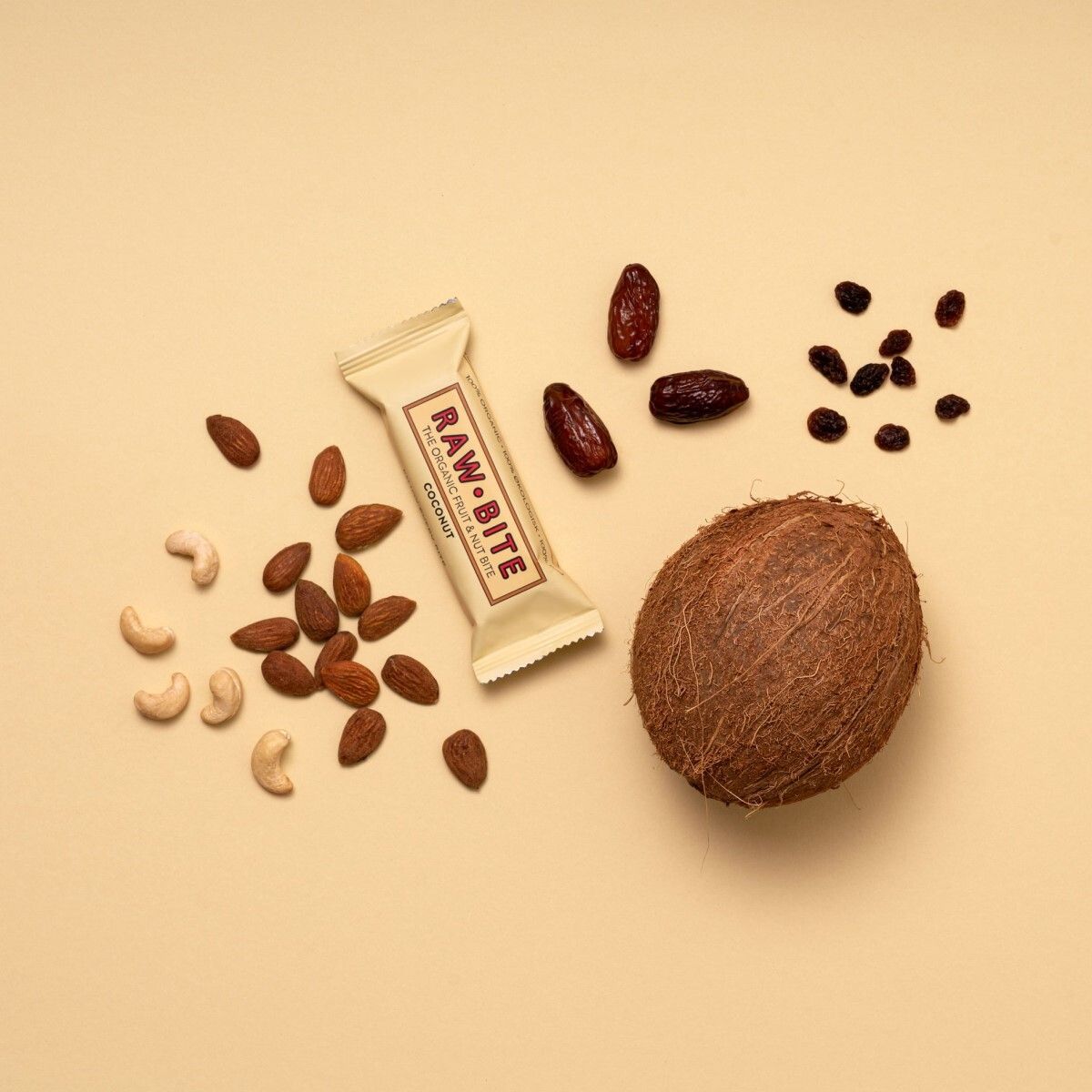 Barre Raw Bite Kokusnuss à côté de noix de coco, amandes, noix de cajou, dattes et raisins secs. Inscription : Raw Bite, noix de coco.