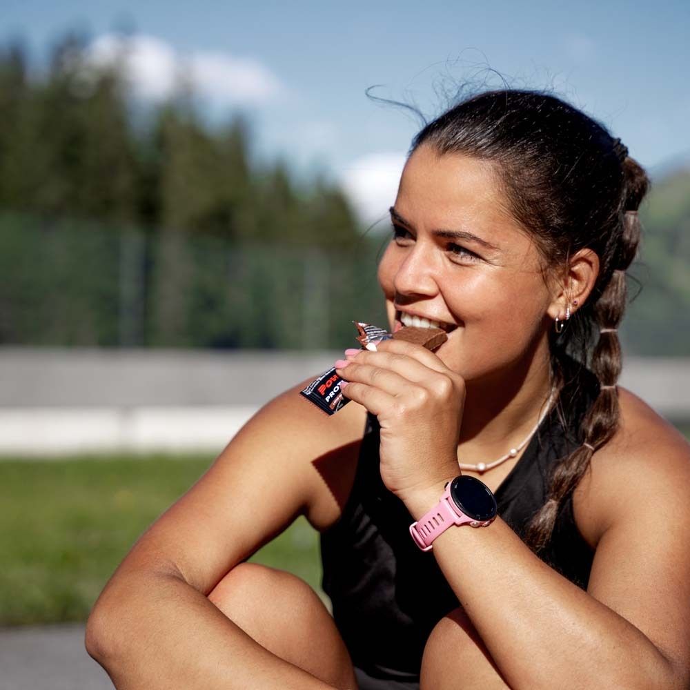 Femme mange une barre Powerbar Protein+. Assise à l'extérieur. Porte une montre intelligente.