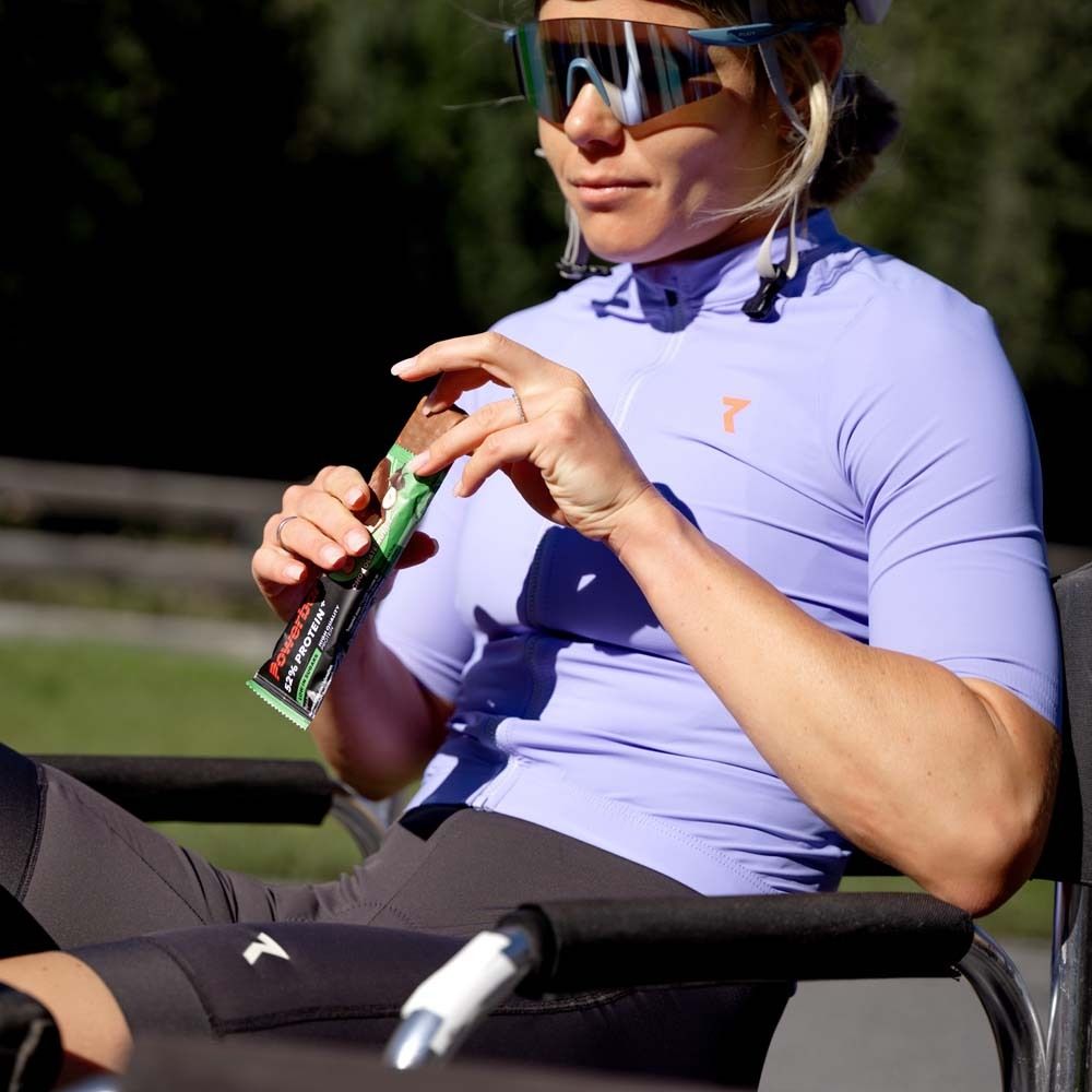 Personne tenant une barre Powerbar 52% Protein Plus. Emballage vert et blanc. La personne est en fauteuil roulant. Nom du produit, emballage.