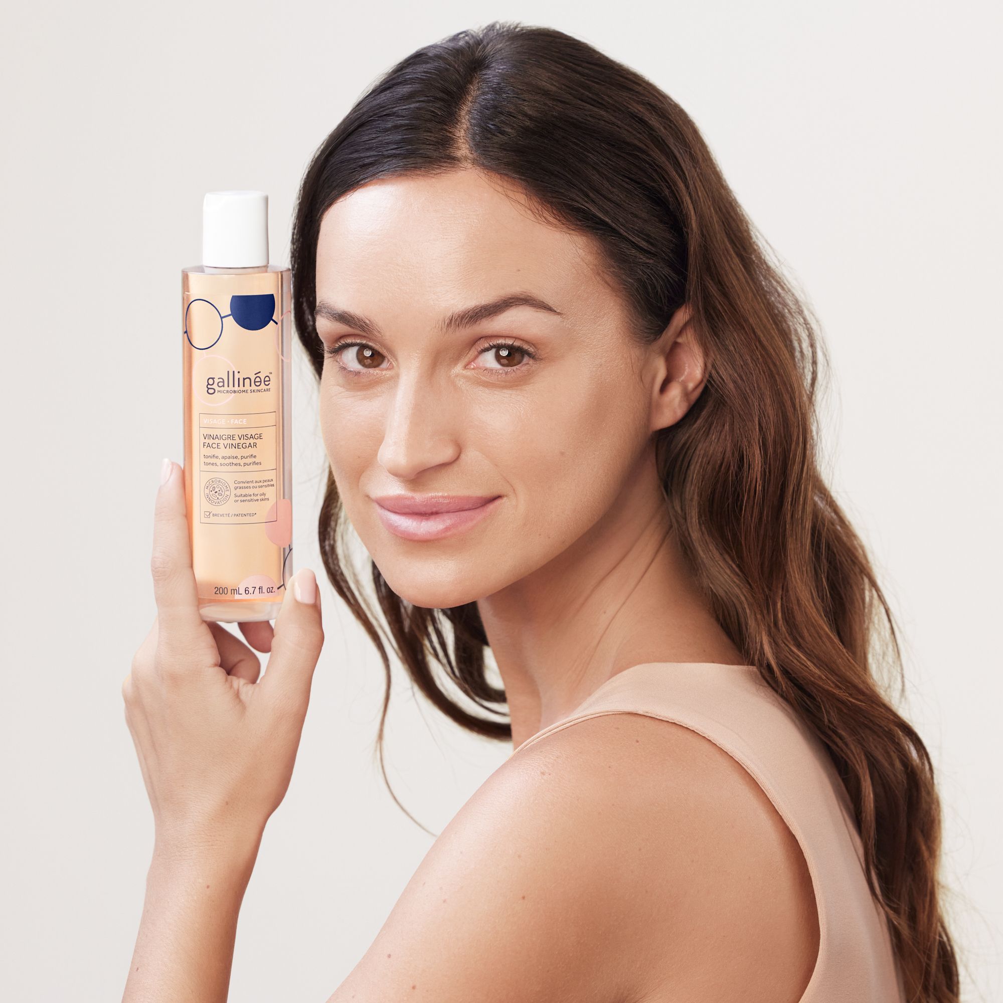 Femme tenant un flacon de Face Vinegar. Cheveux bruns, haut beige. Flacon avec texte.