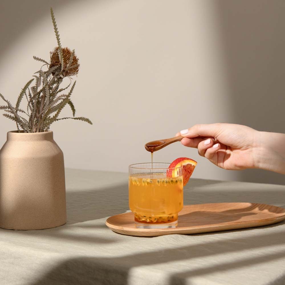 Verre avec boisson, plateau en bois, main avec cuillère, vase avec branche.