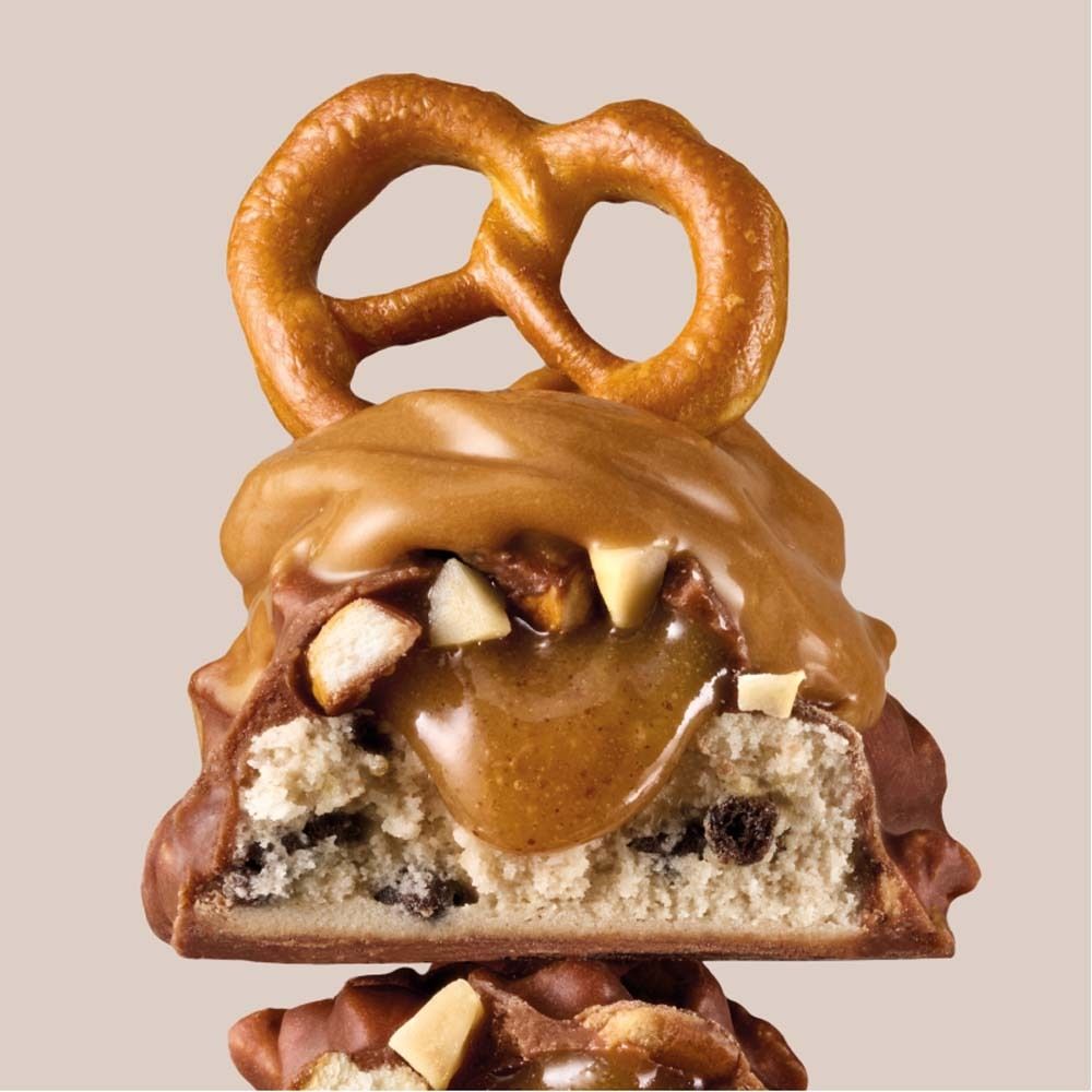 Coupe transversale d'une barre protéinée avec cacahuète, bretzel et caramel. Ingrédients visibles et enrobage chocolat.