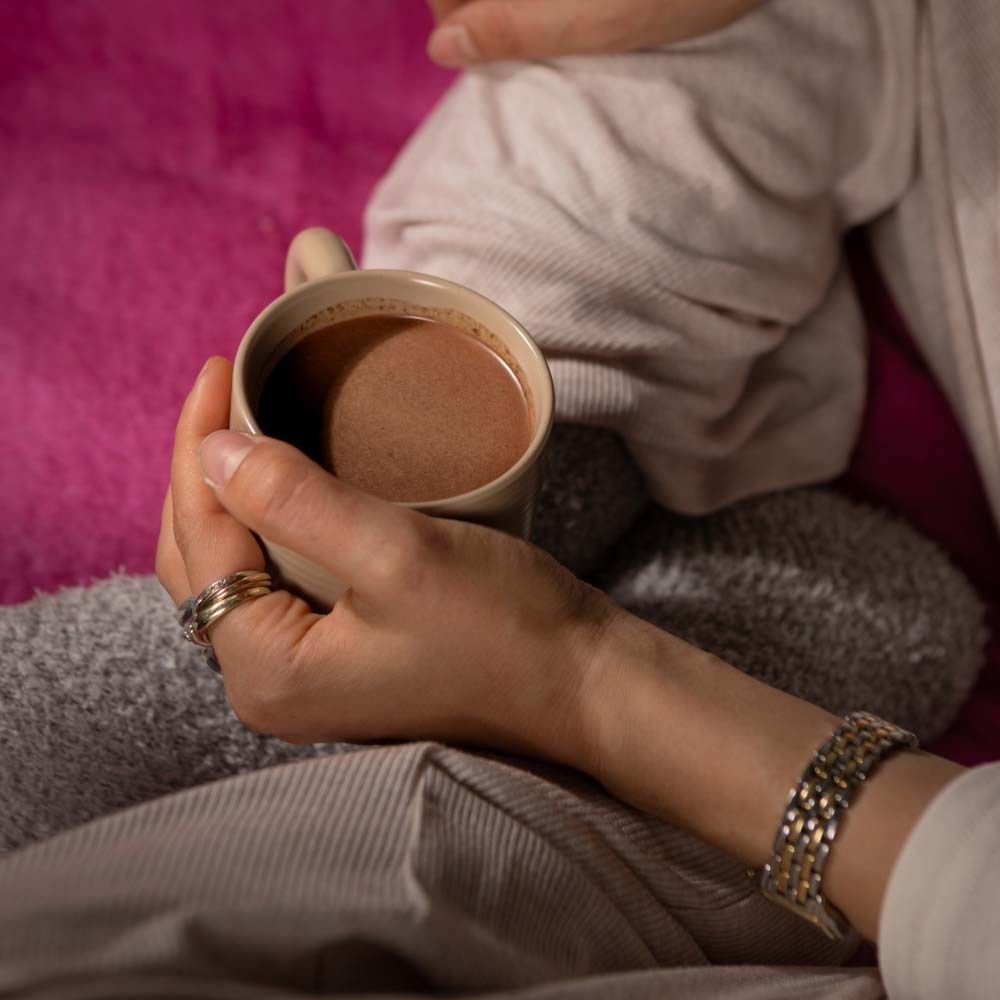 Mains tenant une tasse de cacao. La personne porte des bagues et un bracelet. Fond : couverture rose.