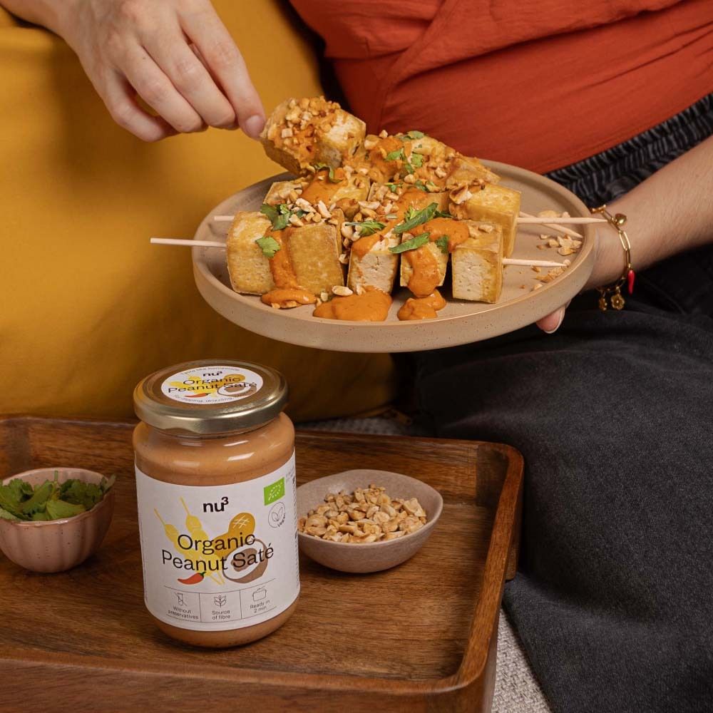 Personne tenant une brochette de tofu et sauce satay aux cacahuètes. À côté, pot de sauce et bol de cacahuètes. Inscription : Organic Peanut Saté.
