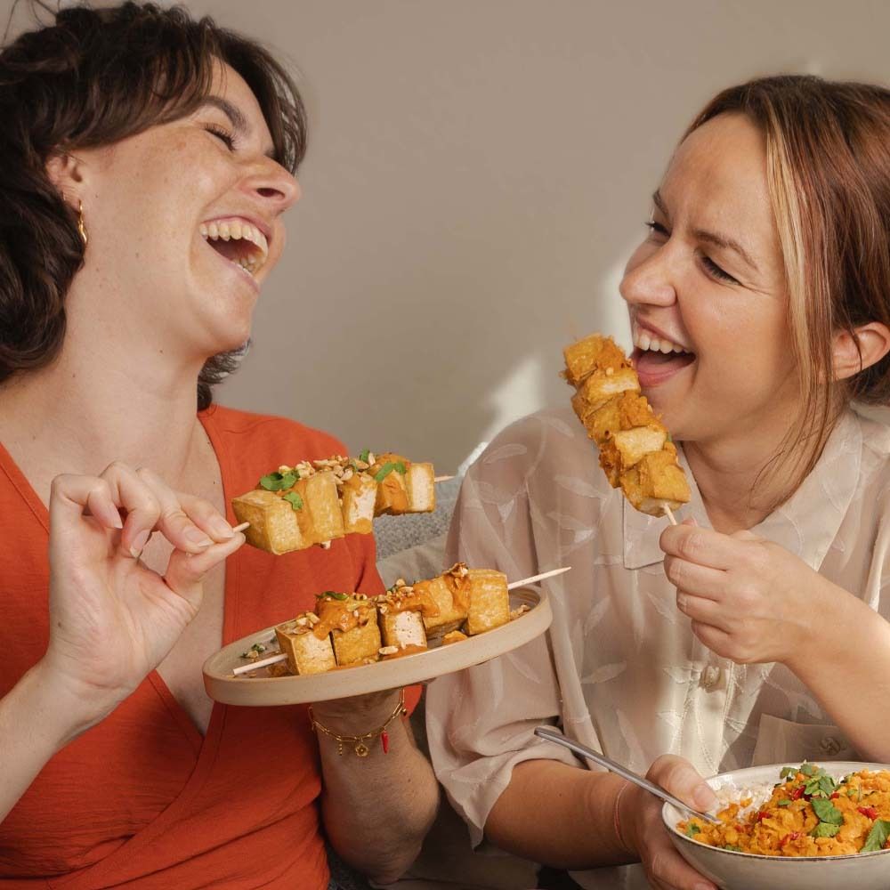 Deux femmes souriantes mangent des brochettes de tofu avec de la sauce satay aux cacahuètes. Sur la table, des bols et un pot de sauce.