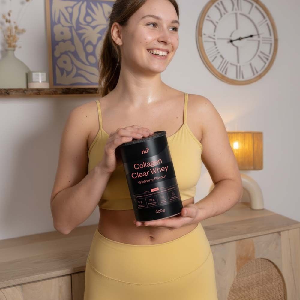Femme tenant une boîte noire nu3 Collagen Clear Whey. Tenue de sport jaune. Arrière-plan : pièce avec horloge et meubles.