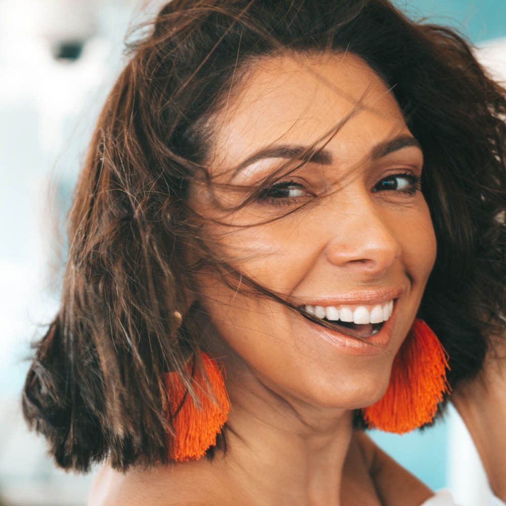 Gros plan d'une femme souriante aux cheveux bruns et aux boucles d'oreilles orange. Elle regarde la caméra.