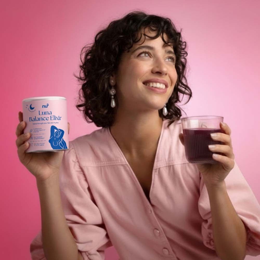 Femme tenant une boîte de nu3 Luna Balance Elixir et un verre de boisson. Fond rose. La femme sourit et regarde vers le haut.