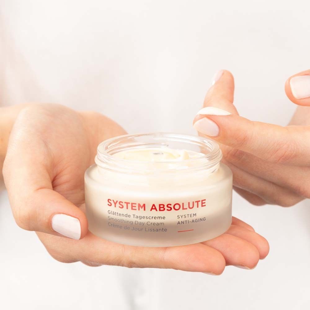 Mains tenant un pot de crème. Inscription : SYSTEM ABSOLUTE, Crème de Jour Lissante, Smoothing Day Cream, Glättende Tagescreme.